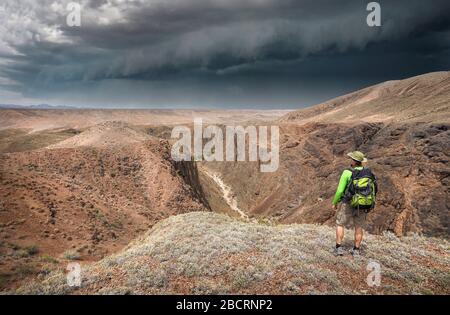 Man with travel backpack in hat and green shirt on top of the canyon looking to the view of valley and storm dark sky Stock Photo