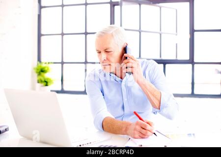 Shot of senior businessman wearing shirt while sitting behind his notebook and making a phone call in the office. Stock Photo