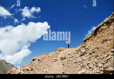 Hiker in yellow shirt going on the trail with rocky mountains at blue cloudy sky background Stock Photo