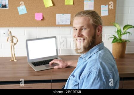 Smiling millennial man looking at camera working from home office on laptop. Stock Photo