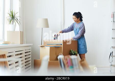 Side view portrait of young Asian woman packing cardboard boxes and smiling happily while moving to new home or apartment, copy space Stock Photo