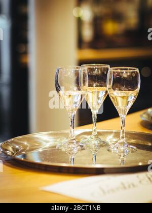 Interior shot of three glasses of white wine on a silver metal tray placed on the table with blurred background in artificial lightning. Stock Photo
