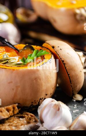 Pumpkin shell with homemade rustic pumpkin soup with seeds on metal tray with garlic and bread aside Stock Photo