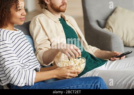 Cropped portrait of modern mixed-race couple watching TV at home while relaxing on cozy sofa, focus on hands holding bowl of popcorn, copy space Stock Photo