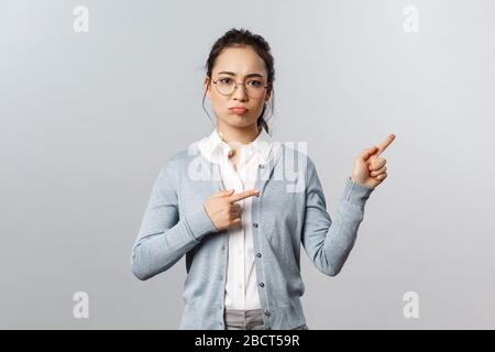 Unamused, gloomy and complaining young asian girl in glasses, sulking upset, grimacing unsatisfied and pointing fingers right with regret and Stock Photo