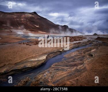 dramatic dark picture of couple walking along geothermal stream with mountains at background against cloudy sky in Hverir, Iceland Stock Photo