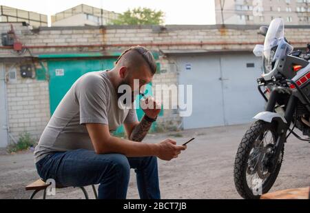 Thoughtful man having rest near bike. Male biker smokes near a bike. A biker looks at his smartphone and smokes an electronic cigarette. Stock Photo
