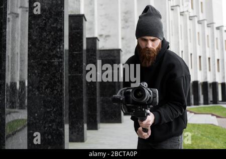 Bearded Professional videographer in black hoodie holding professional camera on 3-axis gimbal stabilizer. Filmmaker making a great video with a Stock Photo