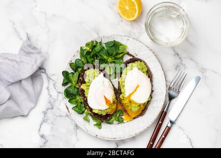 Tasty Healthy Breakfast Toast With Avocado And Poached Egg And Glass Of Pure Water On White Plate Over Marble Background. Table Top View Stock Photo