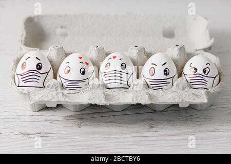 Five white eggs in tray close up with drawn emotional eyes, hearts and medical mask as canceled Easter holiday during april coronavirus quarantine Stock Photo