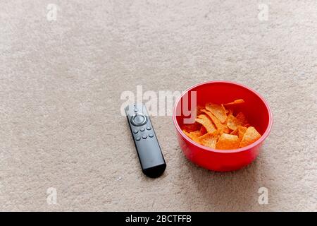 a close up view of a smart tv remote next to a red plastic bowl of chips or crisps on the light coloured carpet Stock Photo