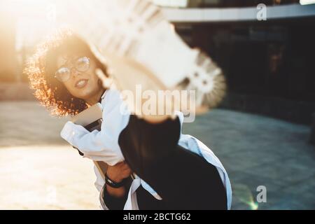Curly haired businesswoman is kicking at camera while posing with some gadgets in eyeglasses outside Stock Photo