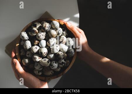 Woman hands holding wooden bowl with quail eggs. Dark background, beautiful natural sun light in kitchen. Easter cooking concept. Stock Photo