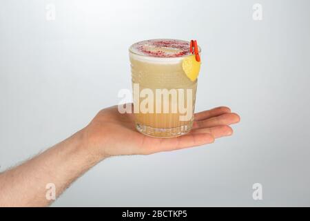 Close up of a hand holding a glass of whiskey sour cocktail on white background with copy space. Stock Photo