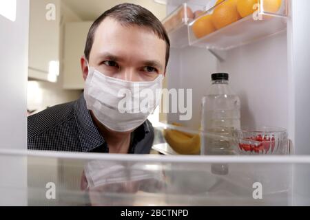 Man in a medical mask looks into the fridge with food. Concept of diet, obesity during home quarantine at covid-19 coronavirus pandemic Stock Photo