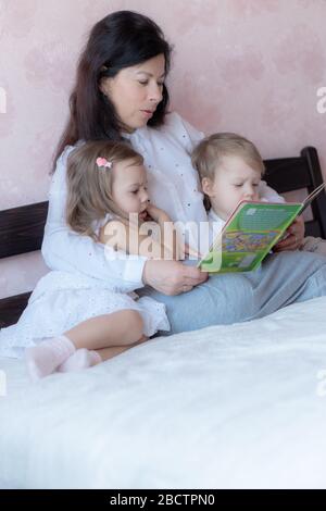 Grandmother with grandson and granddaughter in bed reading a book. Grandmother with grandchildren in bed during distance learning. Grandma plays with Stock Photo