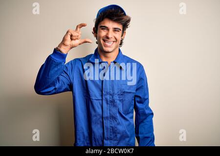 Young mechanic man wearing blue cap and uniform standing over isolated white background smiling and confident gesturing with hand doing small size sig Stock Photo