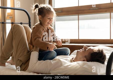 Happy young father lying on bed, holding daughter on belly. Stock Photo