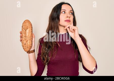 Young beautiful woman holding wholemeal bread cereal over isolated white background serious face thinking about question, very confused idea Stock Photo