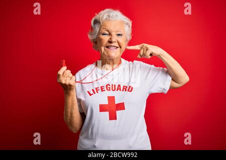Senior beautiful grey-haired lifeguard woman wearing t-shirt with red cross using whistle smiling cheerful showing and pointing with fingers teeth and Stock Photo