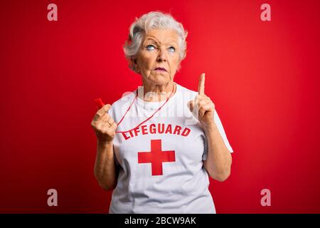 Senior beautiful grey-haired lifeguard woman wearing t-shirt with red cross using whistle Pointing up looking sad and upset, indicating direction with Stock Photo