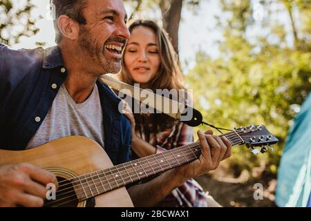 Smiling mature man playing guitar with a woman sitting by. Happy mature couple camping in nature. Stock Photo