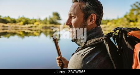 Closeup of a mature man with backpack standing by the lake. Male camper looking at the view. Stock Photo