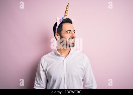Young handsome man with beard wearing funny unicorn diadem over pink background looking away to side with smile on face, natural expression. Laughing Stock Photo