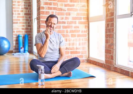 Middle age handsome sportman sitting on mat doing stretching yoga exercise at gym touching mouth with hand with painful expression because of toothach Stock Photo