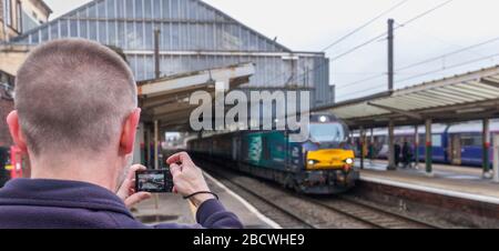 Rail enthusiast / train spotter photographing DRS class 68 68017 Hornet at Preston railway station departing with a Northern Rail passenger service Stock Photo