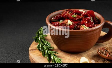 Sun-dried tomatoes with garlic, rosemary and spices in a clay bowl on an olive wood cutting board, copyspace. Stock Photo