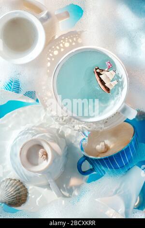 Doing chores concept, toy ship in a coffee cup, white porcelain plates and cups with bubbles and foam Stock Photo