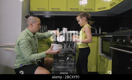 Father teaching daughter smart girl learning to use dishwasher. Young mistress children loading putting dirty dishes in automatic dishwasher.