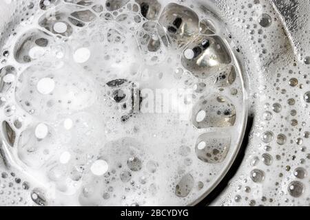 White foam with bubbles of cleaner in a washbasin, macro view. Drain hole with soap bubbles in metal sink. Mechanically adjustable drain plug closeup. Stock Photo