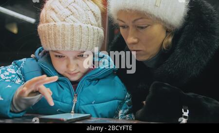 Attractive caucasian mother and daughter using smartphone sitting in cafe. Advanced little child shows and explains to mom something on smartphone. Stock Photo