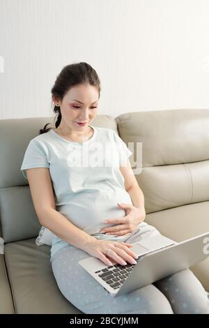 Young pregnant Asian woman in comfortable pajamas sitting on sofa at home watching webinar online Stock Photo