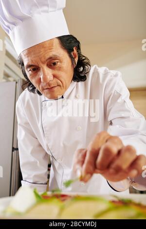 Mature Vietnamese man in chef coat and hat garnishing dish with greens Stock Photo