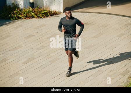 Serious young African-American sportsman running outdoors in shorts and rashguard when preparing for marathone Stock Photo