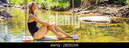 Beautiful happy girl, young pretty cheerful woman traveler sitting, swinging on a swing tied to a tree above the water, smiling in sunny day. Summer Stock Photo