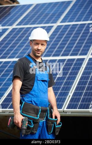 Portrait of happy engineer technician in white protective helmet and blue overalls with electrical screwdriver on background of exterior solar panel photo voltaic system blue shiny surface. Stock Photo
