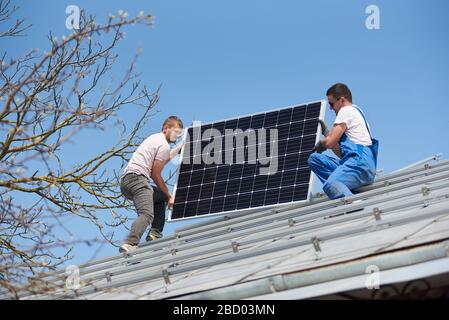 Male engineers installing solar photovoltaic panel system. Two electricians mounting blue solar module on roof of modern house. Alternative energy sustainable resources renewable concept. Stock Photo