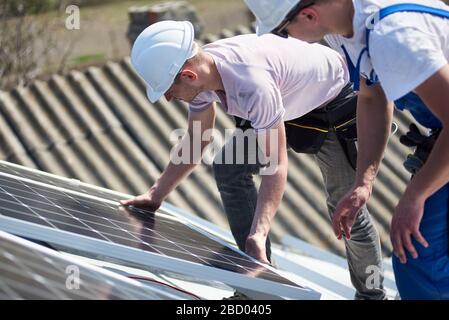 Male engineers installing stand-alone solar photovoltaic panel system. Two electricians mounting blue solar module on roof of modern house. Alternative energy environmental concept. Stock Photo