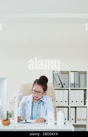 Serious mature female doctor sitting at office desk and taking notes in planner Stock Photo