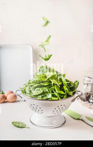 Colander with spinach and falling spinach leaves on white kitchen table. Healthy food concept Stock Photo