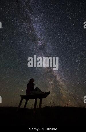 Dark silhouette of female hiker resting at summer night camping in the mountains. Young woman sitting on a bench, enjoying view of night sky full of stars and Milky way. Astrophotography Stock Photo