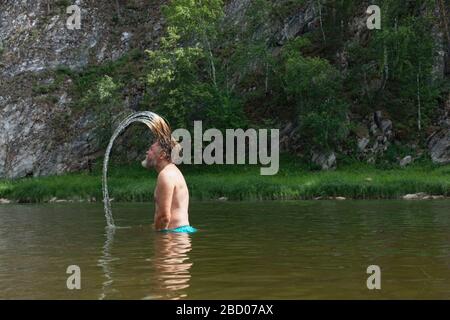 Side view of old man standing waist-deep in the mountain river and throwing his long wet hair back. Tourism, active leisure, bath, soft and silky hair Stock Photo