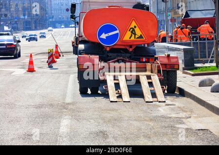 An old trailer with a low platform for transporting road equipment is attached to the rear bumper of an old water carrier and stands against a city. Stock Photo