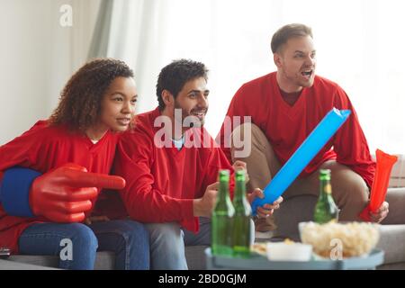 Group of friends watching sports match on TV at home and cheering emotionally while wearing red team uniforms, copy space Stock Photo