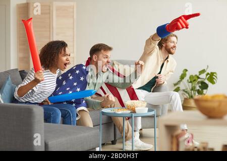 Side view at group of young people watching sports match on TV at home and cheering emotionally while wearing American flag, copy space Stock Photo
