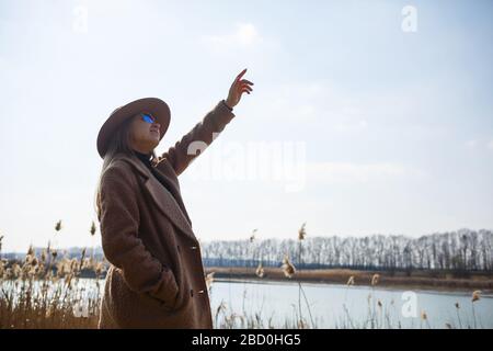 A girl in a brown coat, hat and glasses walks in a park with a lake under the bright sun. Rejoices in life and smiles. The beginning of spring Stock Photo